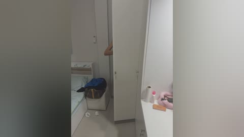 Media: A video of a narrow, cluttered bathroom with white walls and a beige sink counter. A white cabinet and a blue bag are on the floor, and toiletries are scattered on the counter.