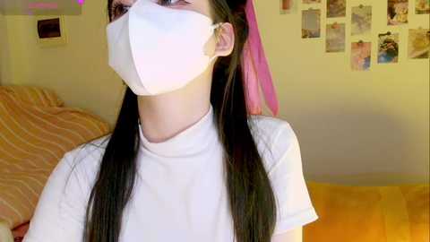 Media: Video of a young woman with long black hair, wearing a white face mask, white T-shirt, and pink headband, standing indoors with a bed, wall, and photos in the background.