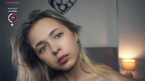 Media: Video of a young, light-skinned woman with long, straight blonde hair and green eyes, wearing no top, looking sad. Background features a dimly lit room with a candle and a clock.