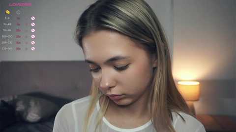 Media: Video of a young, fair-skinned woman with long, straight blonde hair, wearing a white top, looking down with a serious expression. Background features a soft-lit lamp, gray wall, and a blurred bed.