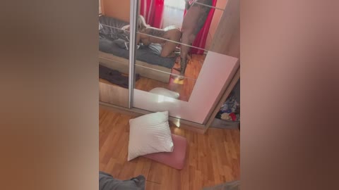 Media: Video of a cozy, cluttered bedroom with wooden floors, a large mirror, a bed with red curtains, and a pink pillow on the floor.