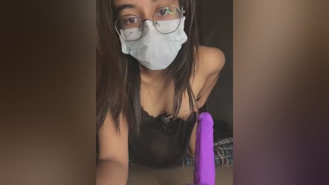 Media: A video of a young woman with long, straight brown hair, wearing a black lace bra, a white surgical mask, and round glasses, leaning over a table with a purple vibrator in her hand.