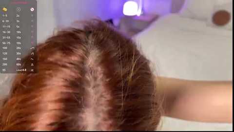 Media: A close-up video of a person's head, focusing on their brown, messy hair, with a digital screen displaying a calendar in the upper left corner. The background features a blurred, softly lit room with purple and white lights.