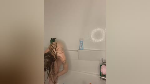 Media: A video of a woman with wet, dark hair, bending over in a white-tiled shower, holding a shampoo bottle, with a pink loofah and a soap dispenser visible.