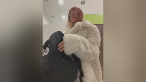 Media: A video of a woman with long, straight, light brown hair, wearing a fluffy white coat and glasses, carrying a black backpack. The background features a beige wall with a green stripe and recessed lighting.