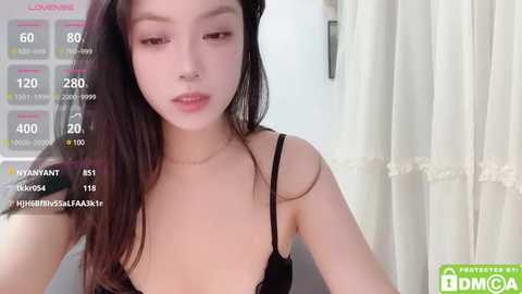 Media: Video of an Asian woman with long black hair, fair skin, and slender build, wearing a black lace bra, taken indoors with a white curtain in the background.