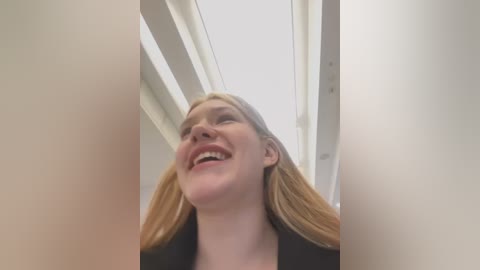 Media: Video of a young woman with long, blonde hair, wearing a black top, smiling from a low angle, in a modern, brightly lit room with white walls and rectangular skylights.