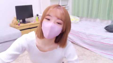 Media: Video of a young Asian woman with shoulder-length light brown hair, wearing a white long-sleeved top and a pink mask, sitting on a beige carpet in a brightly lit bedroom with a white bed, green curtains, and a TV stand.