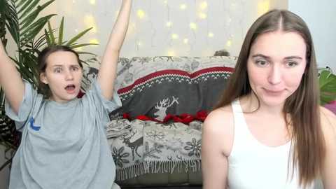 Media: Video of two young women, one with long brown hair, wearing a white tank top, and the other with short brown hair, wearing a gray T-shirt, both excitedly dancing in a cozy living room with a festive throw blanket and string lights.