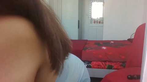 Media: Video of a woman's bare back with shoulder-length brown hair, viewed from behind. She is standing in a small, brightly lit room with a red sofa and a bed with a floral-patterned cover.