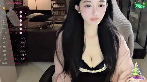 Media: Video of an East Asian woman with long black hair, wearing a black bra and pink cardigan, sitting in a modern living room.