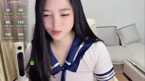 Media: Video of an East Asian woman with long black hair, fair skin, and a petite build wearing a white sailor school uniform, sitting in a modern living room with a gray sectional sofa.