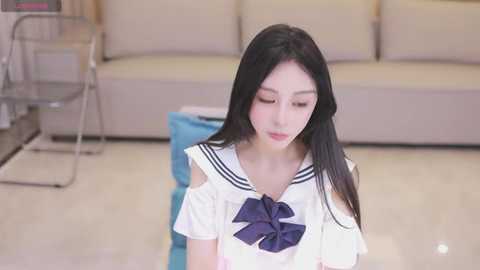 Media: Video of a young East Asian woman with long black hair, wearing a sailor schoolgirl outfit, kneeling on a shiny beige floor in a modern, minimalist living room with beige couches and a blue ottoman.