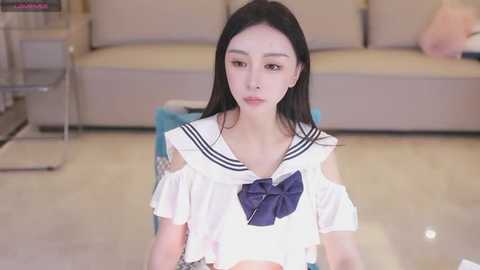 Media: A video of a young East Asian woman with long black hair, fair skin, and a slender physique, wearing a white sailor-style school uniform with a blue bow. She sits on a light-colored floor, indoors with a beige sofa and modern furniture in the background.