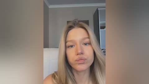 Media: Video of a young woman with long, straight blonde hair, fair skin, and blue eyes, puckering her lips in a playful manner. She wears a sleeveless top. Background shows a beige wall and a partially visible white wardrobe.