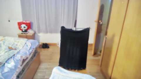 Media: Video of a bedroom with a black umbrella partially open, wooden floor, light-colored walls, a bed with blue and white bedding, a small red radio, and a wooden wardrobe.