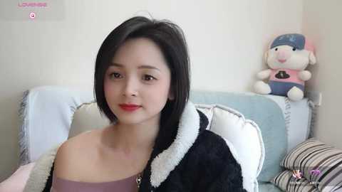 Media: Video of an East Asian woman with straight black hair, fair skin, wearing a purple top and a black and white jacket, seated on a light blue couch.