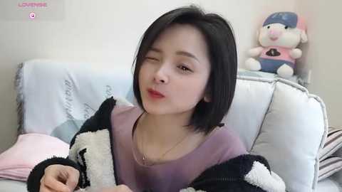 Media: A video of an East Asian woman with straight black hair, wearing a lavender top and a black and white sweater, winking, sitting on a white couch. A plush toy is beside her.