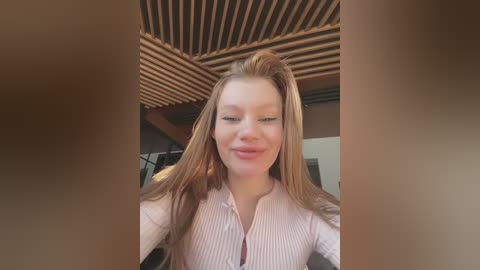 Media: Video of a young, fair-skinned woman with long, straight, light brown hair, wearing a light pink, ribbed, long-sleeve top. She has a slight smile. Background includes wooden ceiling beams and blurred indoor setting.