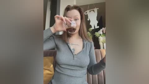 Media: Video of a light-skinned, brunette woman with glasses drinking coffee in a cozy living room. She wears a grey ribbed sweater and has a tattoo on her chest.