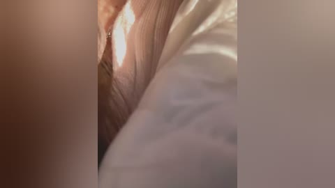 Media: Close-up video of a person's exposed vulva, with light brown skin, smooth texture, and slightly visible pubic hair. The background is blurred, featuring soft, white sheets, creating a private and intimate setting.