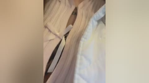 Video of a person's neck and upper chest, wearing a white shirt with a black and white striped tie. The background is blurred and neutral-toned, suggesting an indoor setting.