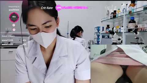 Video of a female Asian lab technician in a white lab coat, mask, and goggles, with a close-up of her exposed buttocks in a pink lab gown. Background: lab equipment, bottles, and a microscope.