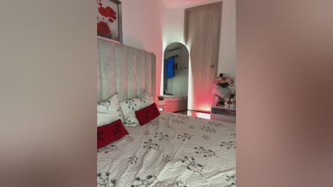 Media: Video of a modern, cozy bedroom with a white bed adorned with red pillows, floral-patterned sheets, and a light grey upholstered headboard. The room features a cream-colored door, a small table with flowers, and a red wall decoration.