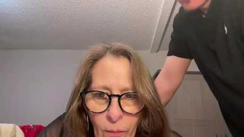 Media: A video of a middle-aged woman with glasses, fair skin, and shoulder-length brown hair, receiving a haircut in a dimly lit room with white walls and a ceiling fan.