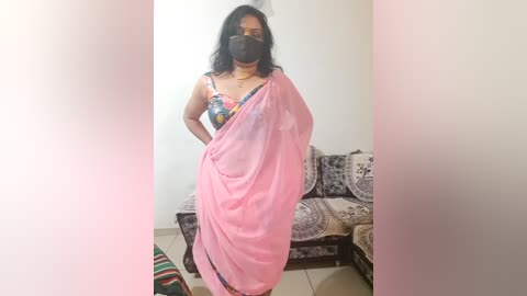 Media: A video of a woman with medium-dark skin, wearing a black mask, floral blouse, and pink saree, standing in a minimalist room with beige walls and a patterned sofa.