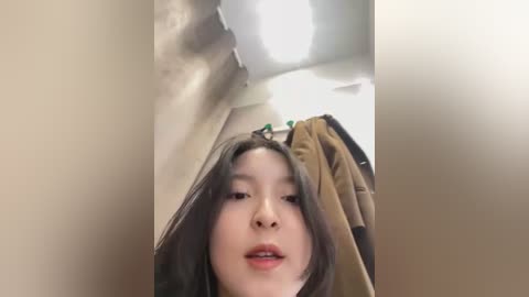 Media: A video of an Asian woman with long black hair, fair skin, and a neutral expression, taken from a low angle. She is wearing a brown jacket, standing in a dimly lit bathroom with beige walls and a ceiling light.