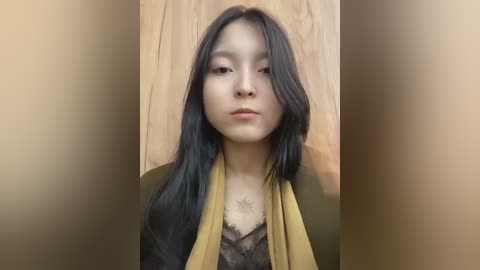 Media: A video of an East Asian woman with long, straight black hair, fair skin, and a neutral expression, wearing a black lace top and mustard-yellow scarf. The background features a beige wall and a wooden door.