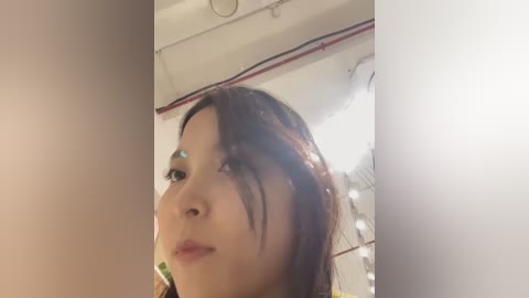 Media: Video of an Asian woman with long black hair and light skin, looking down, in a dimly lit room with white walls and a ceiling light.