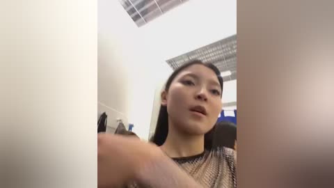 Media: A video of a young Asian woman with straight black hair, wearing a mesh top, standing in an office with fluorescent lights, pointing at a white wall.