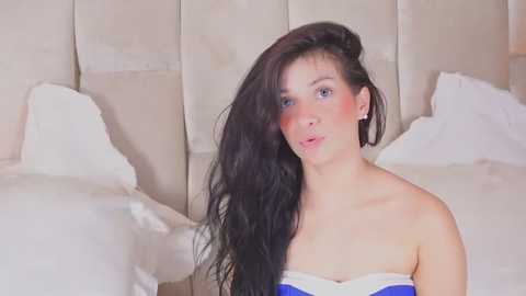 Media: A video of a young Caucasian woman with long, dark hair, wearing a strapless blue and white top, sitting on a bed with white sheets and beige padded headboard.