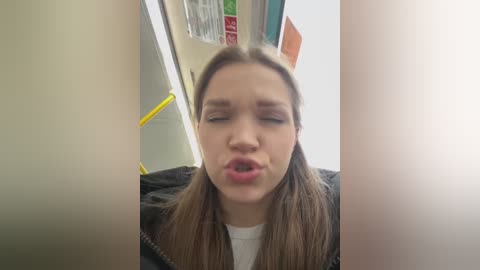 Video of a young woman with straight brown hair and light skin, wearing a black jacket, leaning against a yellow handrail in a subway car. Her eyes are closed, and she is blowing a kiss.