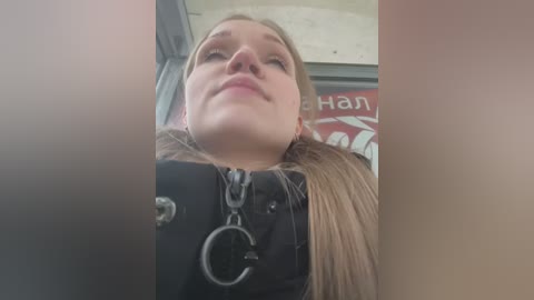 A video of a young woman with fair skin and long blonde hair, wearing a black jacket and a handcuff key necklace, captured from a low angle in a subway station.