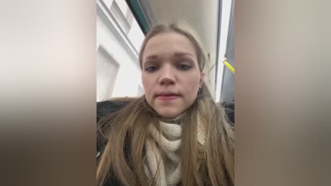 Media: Video of a young woman with light skin and long, straight blonde hair, wearing a beige scarf and black jacket, sitting inside a bus with a blurred background.