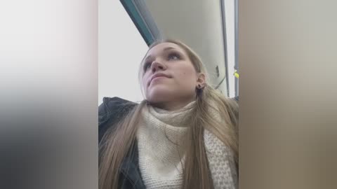 Video of a blonde woman with long hair, wearing a beige scarf and a dark jacket, looking upward, inside a subway train.