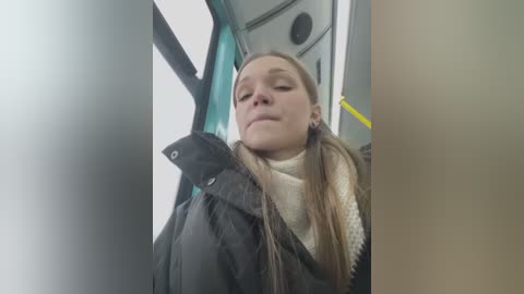 Media: Video of a young Caucasian woman with long brown hair, eyes closed, wearing a black raincoat over a cream sweater, seated in a bus.