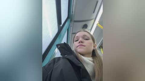 Media: Video of a young woman with fair skin and long blonde hair, wearing a black jacket and beige scarf, sitting inside a modern, clean train with blue and gray interior.