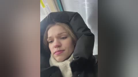 Video of a young Caucasian woman with light skin and blonde hair, wearing a black winter coat and a white scarf, appearing tired and yawning inside a dimly lit room.