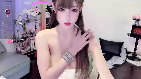 Media: Video of a pale, slender, nude woman with long brown hair, wearing a silver bracelet, sitting on a black cushioned chair in a modern room with a metal bookshelf and pink flowers.