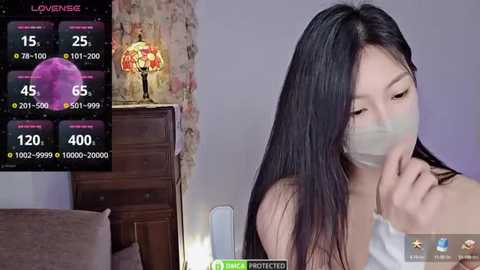 Media: Video of an East Asian woman with long black hair and a surgical mask, standing in a dimly lit room with a wooden dresser, a floral lamp, and a digital display showing COVID-19 statistics.