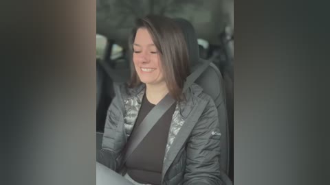 Media: Video of a woman with shoulder-length brown hair, wearing a gray puffy jacket and black top, smiling while seated in a car with a gray seatbelt across her chest.