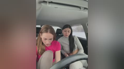 Media: Video of two young women in a car, one with long blonde hair in a pink top, the other with long black hair in a gray sweater, both looking down, car interior visible.