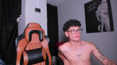 Media: Video of a shirtless, slender, curly-haired young man with glasses and tattoos, wearing orange and black gaming chair, gray walls, Marilyn Monroe poster, and black curtains.