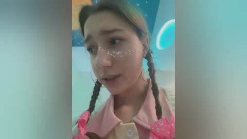 Media: Video of a young woman with pale skin, brown hair in braids, wearing a pink collared shirt, and a pink bow on her head, with white face paint, standing in a room with a blue and white theme.