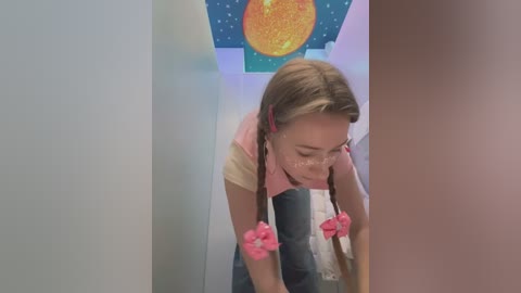 Media: Video of a young girl with long brown hair, wearing a pink shirt and blue jeans, looking down, standing in a hallway with a sun and moon mural on the wall.