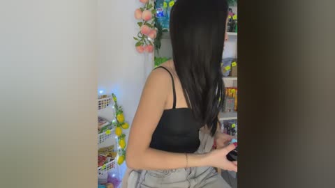 A video of a young woman with straight, black hair, wearing a black tank top and gray shorts, playing a video game in a cluttered room with pastel-colored decorations and shelves filled with toys and games.